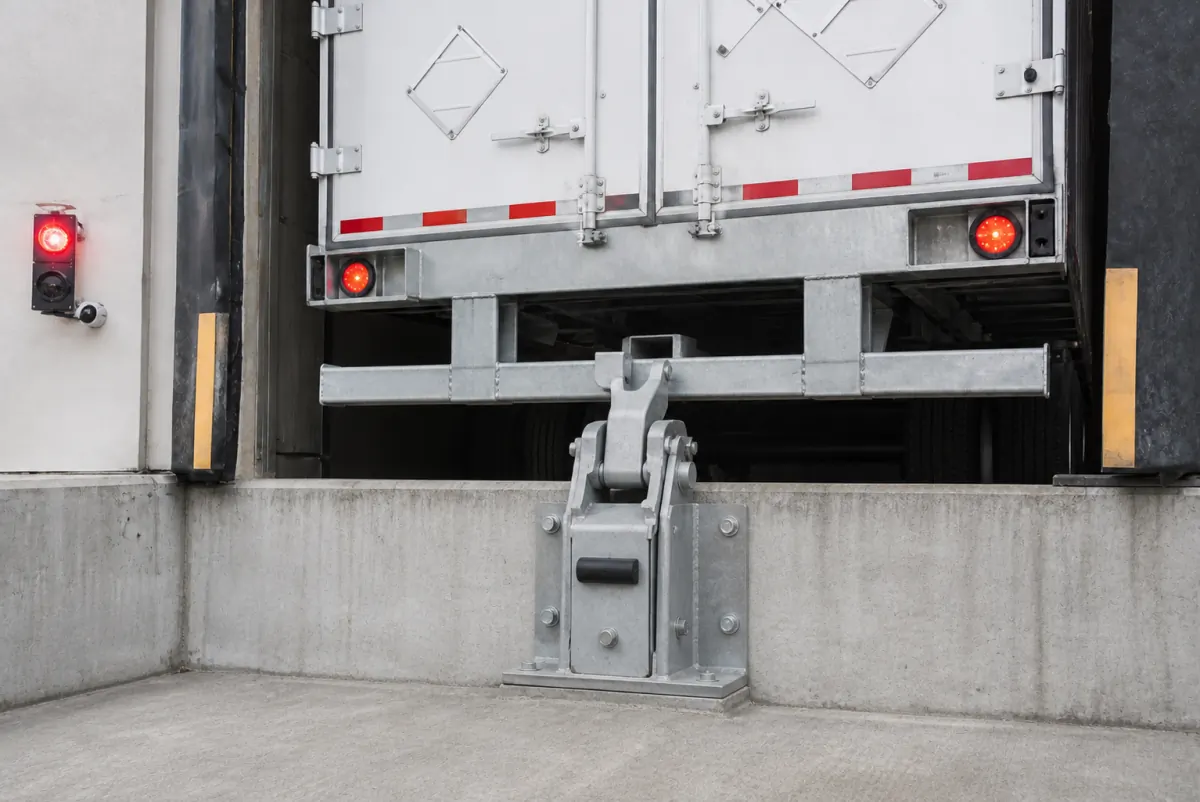 Galvanized vehicle restraint engaged on a trailer's rear impact guard at a loading dock, with a red exterior signal light showing the trailer is locked